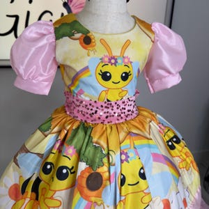 K&ouml;nnte beinhalten: Ein gelbes und rosa Kleid mit einem Bienenmotiv. Das Kleid zeigt eine Cartoon-Biene, Regenbogen und Blumen. Es hat rosa Puff&auml;rmel und einen paillettenbesetzten G&uuml;rtel. Der Rock hat ein farbenfrohes Muster mit Bienen und Sonnenblumen.