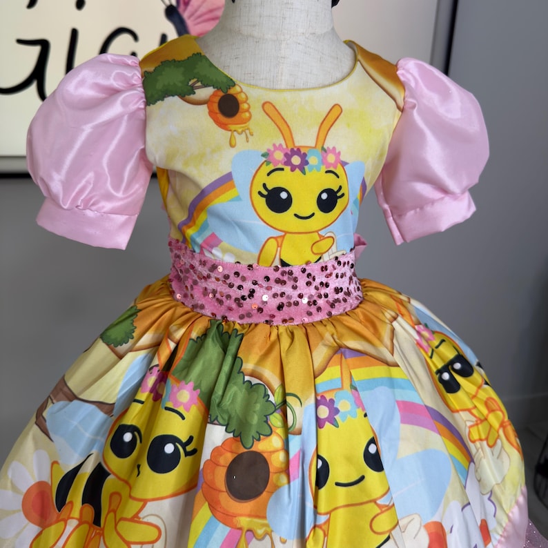 K&ouml;nnte beinhalten: Ein gelbes und rosa Kleid mit einem Bienenmotiv. Das Kleid zeigt eine Cartoon-Biene, Regenbogen und Blumen. Es hat rosa Puff&auml;rmel und einen paillettenbesetzten G&uuml;rtel. Der Rock hat ein farbenfrohes Muster mit Bienen und Sonnenblumen.