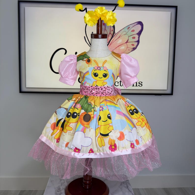 K&ouml;nnte beinhalten: Ein gelb-rosa Kleid im Bienen-Thema mit passendem Stirnband und Schmetterlingsfl&uuml;geln. Das Kleid hat einen Cartoon-Bienen-Print, rosa &Auml;rmel und einen glitzernden rosa T&uuml;llrock. Das Kleid wird auf einer Schaufensterpuppe pr&auml;sentiert.