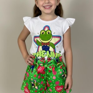 May include: A white t-shirt with a green frog graphic and the word "HENNA" paired with green shorts featuring a frog and heart pattern. The shorts have a large green bow at the waist. A young girl is wearing the outfit.