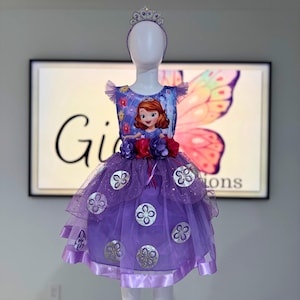 May include: A purple princess dress with a Sofia the First graphic on the bodice. The dress features a layered tulle skirt with silver embellishments, a matching headband, and a tiara. The dress is displayed on a mannequin.