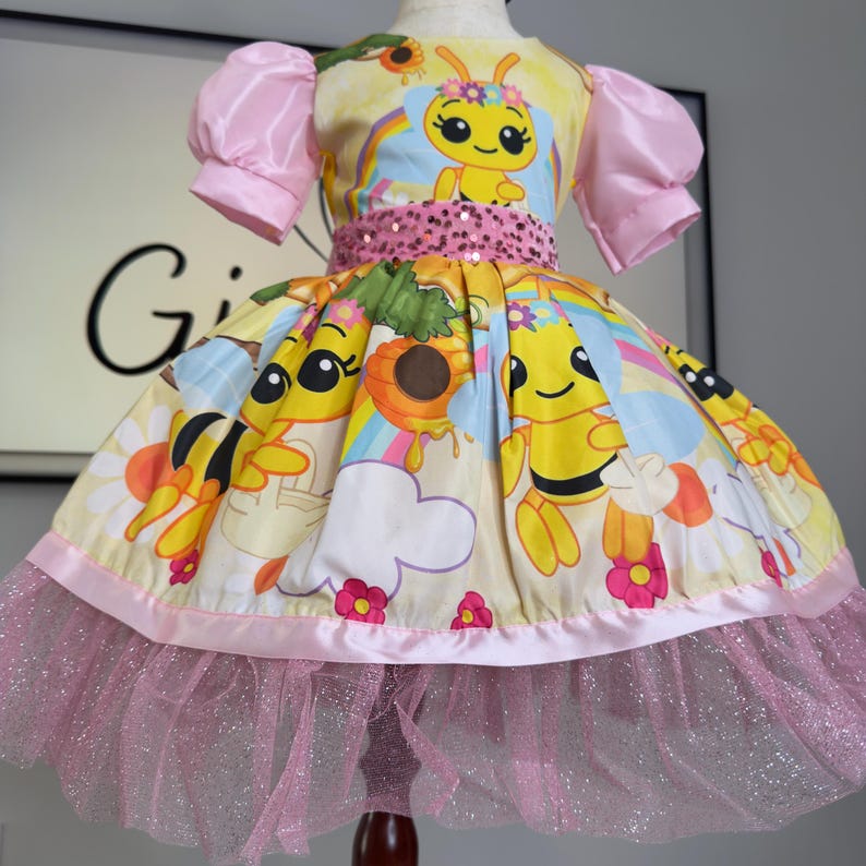 K&ouml;nnte beinhalten: Gelb-rosa Kinderkleid mit Bienen-Motiv. Das Kleid zeigt eine Cartoon-Biene, Regenbogen und Blumen. Es hat rosa Puff&auml;rmel, einen Pailletten-G&uuml;rtel und einen glitzernden T&uuml;llrock.