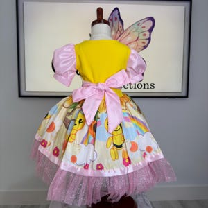 K&ouml;nnte beinhalten: Ein gelbes und rosa Kleid mit einer Schleife auf dem R&uuml;cken. Das Kleid hat Puff&auml;rmel und einen Rock mit einem bunten Aufdruck von Bienen, Regenbogen und Blumen. Der Rock hat einen rosa Glitzersaum.