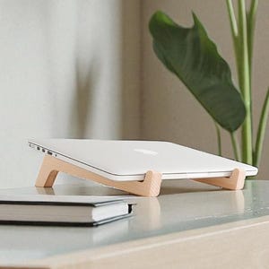 Könnte beinhalten: Ein weißer Laptop steht auf einem Laptop-Ständer aus hellem Holz auf einem hellen Schreibtisch. Ein schwarzes Notizbuch mit weißem Rand befindet sich vor dem Laptop-Ständer. Im Hintergrund befindet sich eine große grüne Pflanze.