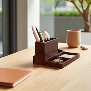 May include: A dark brown wooden desk organiser with a pen holder, tray, and notepad. The organiser holds pens, glasses, and a paper clip. A brown leather-bound notebook and a beige mug are also on the light wood desk.