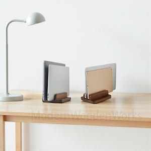 May include: Two wooden laptop and tablet stands on a light wood desk. A silver desk lamp is on the left. The stands are made of dark wood and hold a silver laptop and a gold tablet. The text "Modern Wooden Works" is at the bottom.