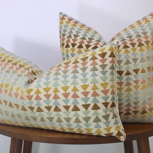 May include: Three decorative throw pillows with a geometric triangle pattern in shades of yellow, brown, pink, and blue. The pillows are on a wooden table, and the background is white.