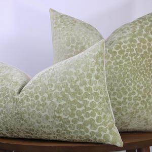 May include: Three decorative throw pillows with a light green, textured, circular pattern. The pillows are arranged on a dark brown wooden table. The pillows have a soft, inviting appearance, perfect for home decor.