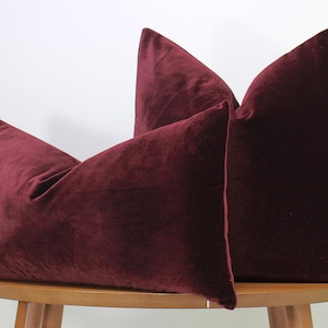 May include: Two burgundy velvet throw pillows are displayed on a wooden table. The pillows have a soft, plush texture and a rich, deep color. The pillows are rectangular and have a classic design, perfect for adding a touch of elegance to any living space.