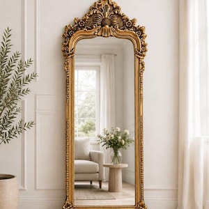 May include: A tall, floor-standing mirror featuring an ornate, gold-colored frame with intricate carved detailing along the arched top and sides. The rectangular glass surface reflects a room interior with a chair, a small table, and a vase.