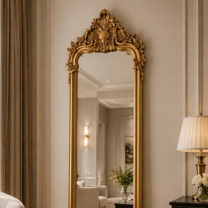May include: A tall, rectangular floor mirror featuring an ornate gold frame with intricate carved floral and scrollwork details at the top and bottom. The mirror has a smooth, reflective glass surface and a classic, arched top design.