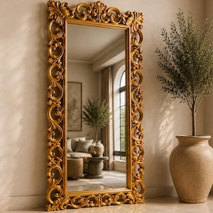 May include: A tall, rectangular floor mirror featuring an ornate, gold-colored frame with intricate scrollwork and carved floral details. The mirror has a reflective glass surface that shows a room interior, and it stands upright against a wall.