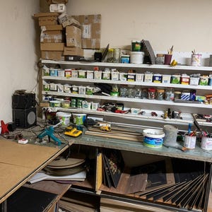 May include: A workshop area featuring a white multi-tiered shelving unit filled with various small containers, jars, and supplies. A red wall clock, a teal glue gun, a yellow power tool, and several paint cans sit on the workbench surface.