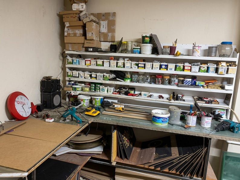 May include: A workshop area featuring a white multi-tiered shelving unit filled with various small containers, jars, and supplies. A red wall clock, a teal glue gun, a yellow power tool, and several paint cans sit on the workbench surface.