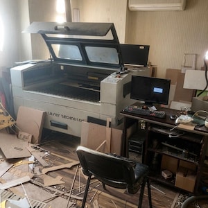 May include: A large industrial laser cutting machine with an open lid, featuring a white exterior and the text Laser Technology on the front. The machine is connected to a computer workstation with a monitor, keyboard, and a black office chair.