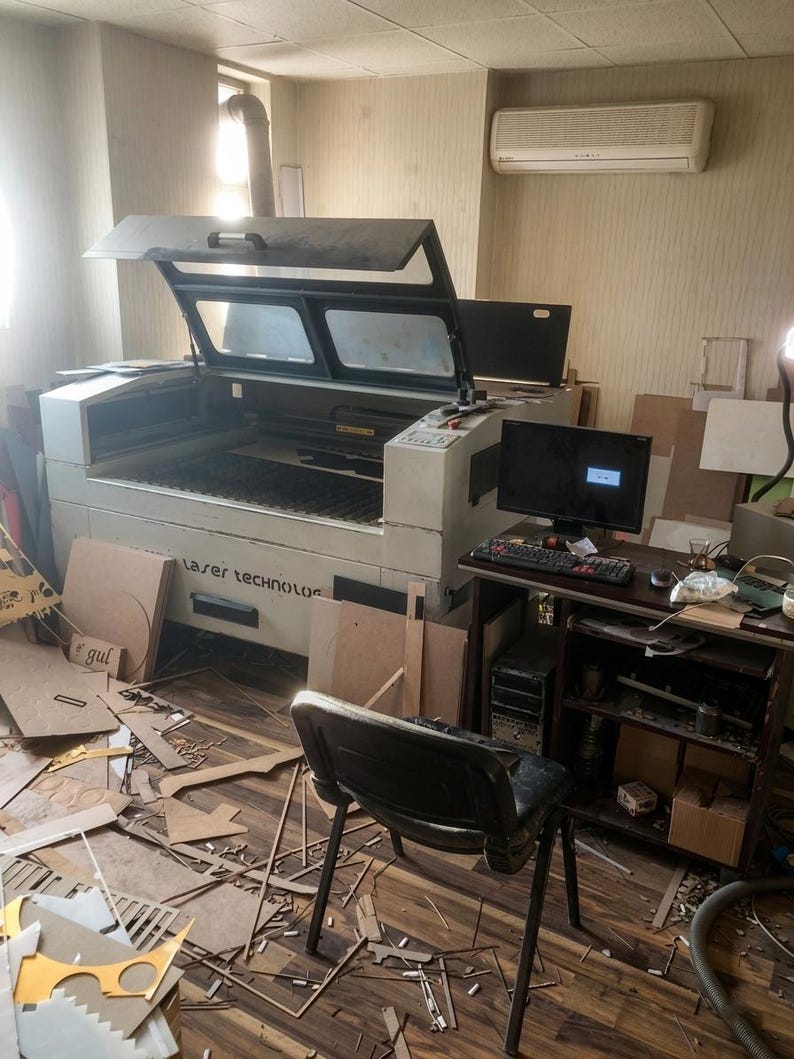 May include: A large industrial laser cutting machine with an open lid, featuring a white exterior and the text Laser Technology on the front. The machine is connected to a computer workstation with a monitor, keyboard, and a black office chair.