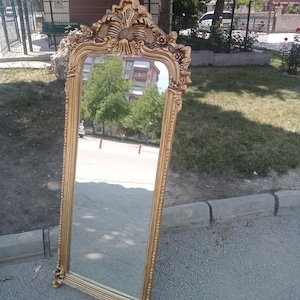 May include: A tall, rectangular floor mirror featuring an ornate, gold-colored frame with intricate carved floral and scrollwork details at the top and bottom. The mirror has a slender, textured border and stands upright on a flat surface.