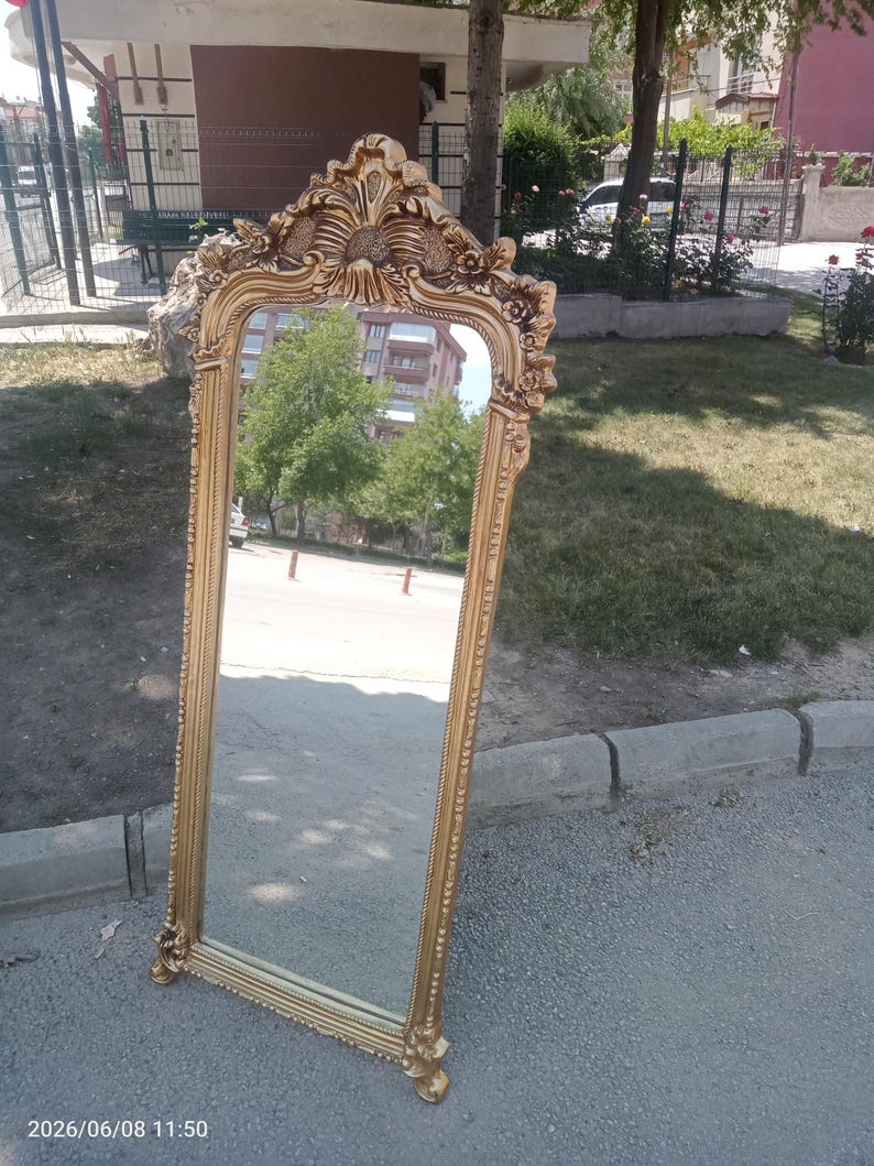 May include: A tall, rectangular floor mirror featuring an ornate, gold-colored frame with intricate carved floral and scrollwork details at the top and bottom. The mirror has a slender, textured border and stands upright on a flat surface.