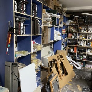 May include: A workshop storage area featuring tall blue shelving units filled with various art supplies, including stacked frames, rolled papers, and containers. A caulking gun hangs on the side, and a square clock with a geometric pattern sits on the floor.