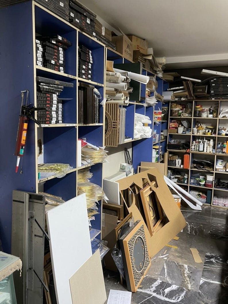 May include: A workshop storage area featuring tall blue shelving units filled with various art supplies, including stacked frames, rolled papers, and containers. A caulking gun hangs on the side, and a square clock with a geometric pattern sits on the floor.