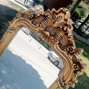 May include: A gold-colored wall mirror featuring an ornate, heavy frame with intricate relief carvings of flowers and textured floral patterns. The frame has a polished finish with deep grooves and raised decorative elements along the edges.