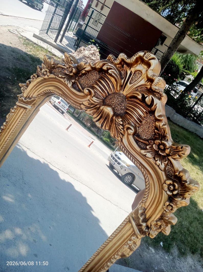 May include: A gold-colored wall mirror featuring an ornate, heavy frame with intricate relief carvings of flowers and textured floral patterns. The frame has a polished finish with deep grooves and raised decorative elements along the edges.