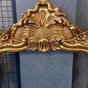 May include: A gold-colored ornate mirror frame featuring intricate relief carvings of floral motifs and a central shell-like crest at the top. The frame has a polished metallic finish with textured detailing throughout the decorative scrollwork.