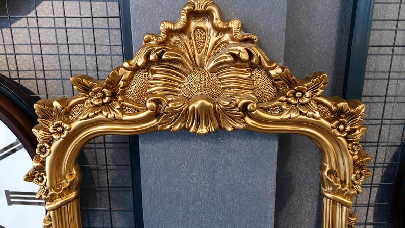 May include: A gold-colored ornate mirror frame featuring intricate relief carvings of floral motifs and a central shell-like crest at the top. The frame has a polished metallic finish with textured detailing throughout the decorative scrollwork.