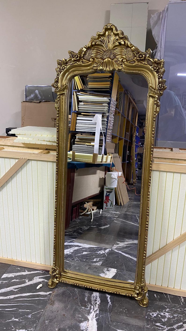 May include: A tall, rectangular floor mirror featuring an ornate, metallic gold frame with intricate carved floral and scrollwork details at the top arch and along the sides. The mirror has a rope-like texture bordering the glass surface.