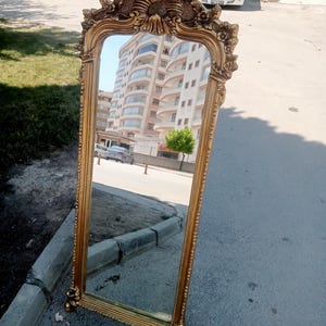 May include: A tall, rectangular floor mirror featuring an ornate, gold-colored frame with intricate floral and scrollwork carvings at the top and bottom. The mirror surface reflects a multi-story residential building and a street scene.