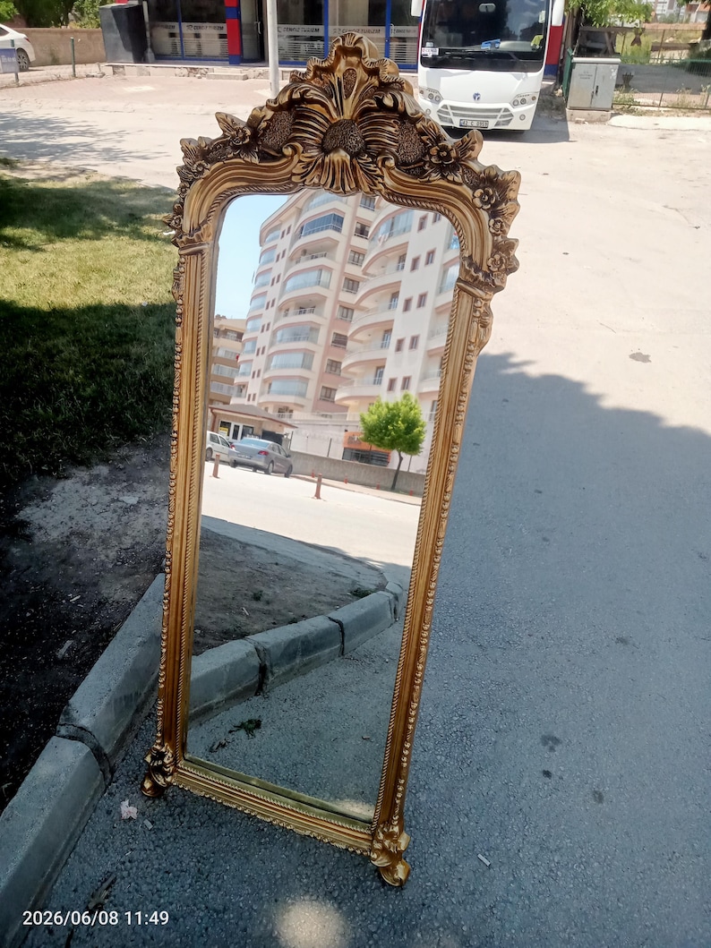 May include: A tall, rectangular floor mirror featuring an ornate, gold-colored frame with intricate floral and scrollwork carvings at the top and bottom. The mirror surface reflects a multi-story residential building and a street scene.