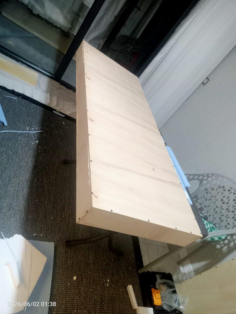 May include: A rectangular, light-colored wooden shelf or panel featuring a smooth, flat surface with visible grain. The side edge is constructed with multiple small, evenly spaced metal fasteners or nails securing the frame together.