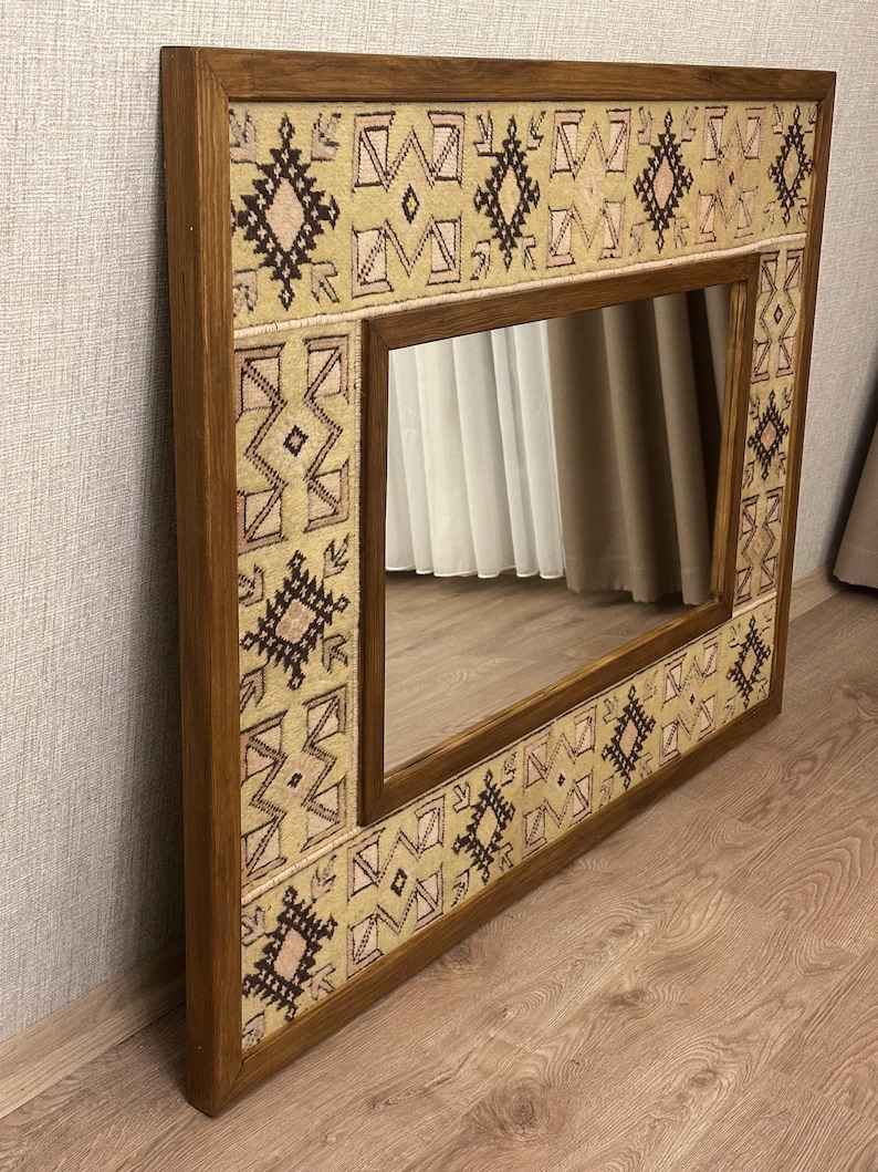 May include: A rectangular mirror with a wooden frame. The frame is adorned with a beige and brown patterned fabric. The mirror reflects the room's interior, showcasing a neutral color palette and adding a decorative element to the space.