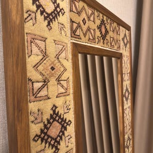 Puede incluir: Un espejo de pared rectangular con un marco de madera en acabado marr&oacute;n medio. El marco est&aacute; incrustado con paneles de tela tejida en tonos beige, rosa claro y marr&oacute;n oscuro, mostrando un patr&oacute;n geom&eacute;trico repetitivo en toda su superficie.