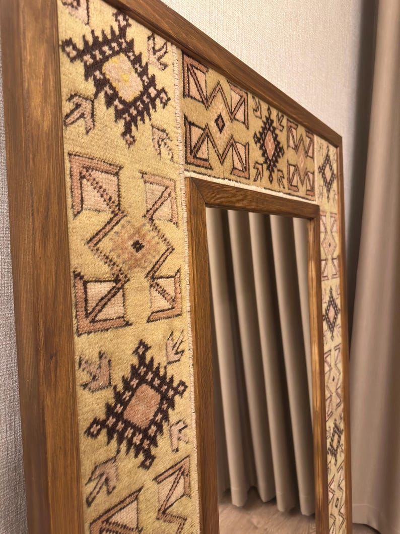Puede incluir: Un espejo de pared rectangular con un marco de madera en acabado marr&oacute;n medio. El marco est&aacute; incrustado con paneles de tela tejida en tonos beige, rosa claro y marr&oacute;n oscuro, mostrando un patr&oacute;n geom&eacute;trico repetitivo en toda su superficie.