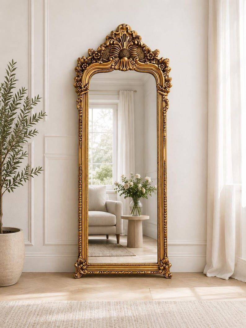 May include: A tall, floor-standing mirror featuring an ornate, gold-colored frame with intricate carved detailing along the arched top and sides. The rectangular glass surface reflects a room interior with a chair, a small table, and a vase.