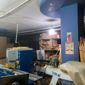 May include: A blue industrial woodworking machine with a yellow and black striped safety guard, a circular saw blade, and a white dust collection unit labeled 2800 m3, situated in a workshop filled with stacks of lumber and a fire extinguisher sign.