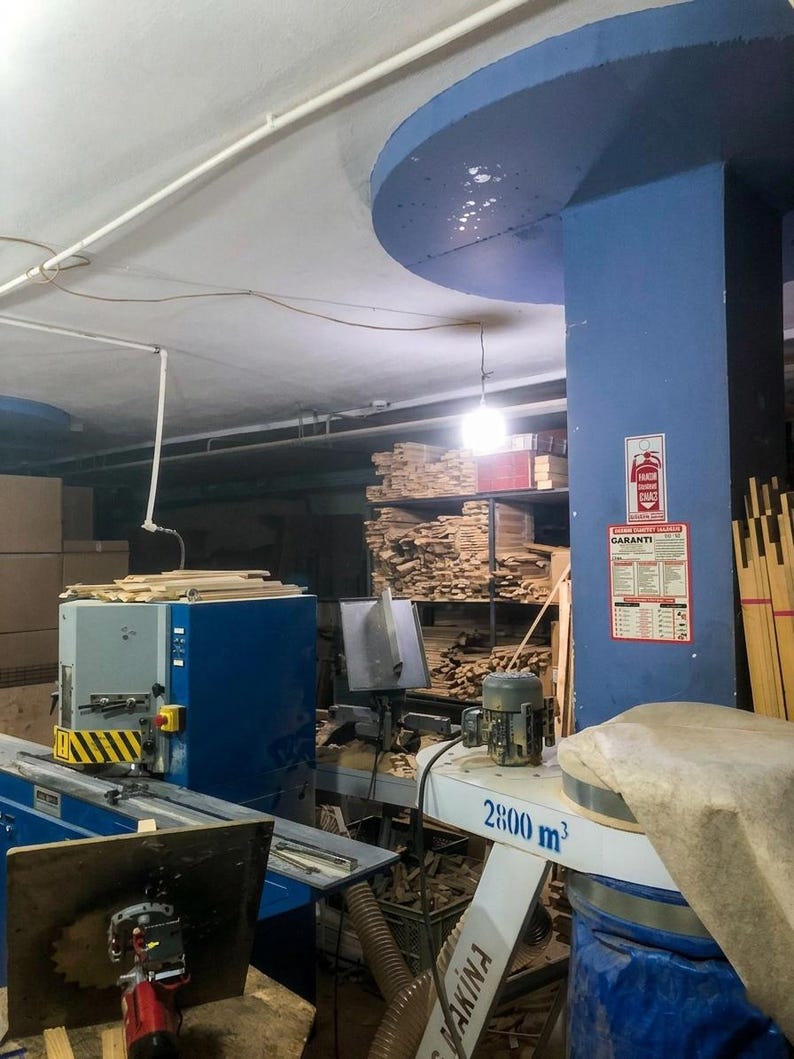 May include: A blue industrial woodworking machine with a yellow and black striped safety guard, a circular saw blade, and a white dust collection unit labeled 2800 m3, situated in a workshop filled with stacks of lumber and a fire extinguisher sign.