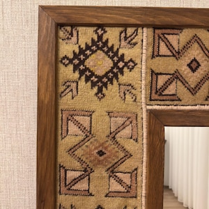 Puede incluir: Un espejo rectangular con un marco de madera de color marr&oacute;n oscuro. El borde interior del marco est&aacute; decorado con un panel de tela tejida texturizada en tonos beige, marr&oacute;n y rosa suave, con patrones geom&eacute;tricos y rombos repetidos.