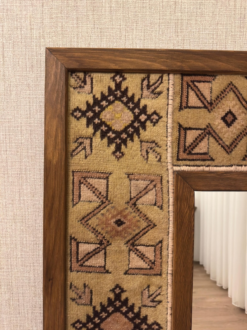 Puede incluir: Un espejo rectangular con un marco de madera de color marr&oacute;n oscuro. El borde interior del marco est&aacute; decorado con un panel de tela tejida texturizada en tonos beige, marr&oacute;n y rosa suave, con patrones geom&eacute;tricos y rombos repetidos.