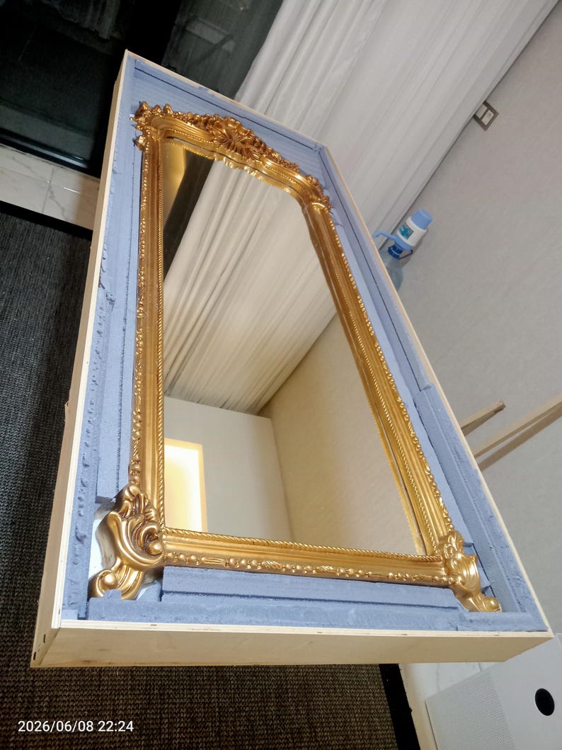 May include: A tall rectangular mirror with an ornate gold-colored frame featuring intricate carved detailing at the corners and edges. The mirror is securely packed inside a light-colored wooden crate lined with blue foam padding for protection.