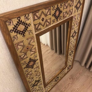 Puede incluir: Un espejo de pared rectangular con un marco de madera de color marr&oacute;n oscuro. El marco est&aacute; incrustado con un borde de tela beige texturizada, decorado con patrones geom&eacute;tricos repetitivos en tonos marr&oacute;n oscuro, negro y rosa suave.
