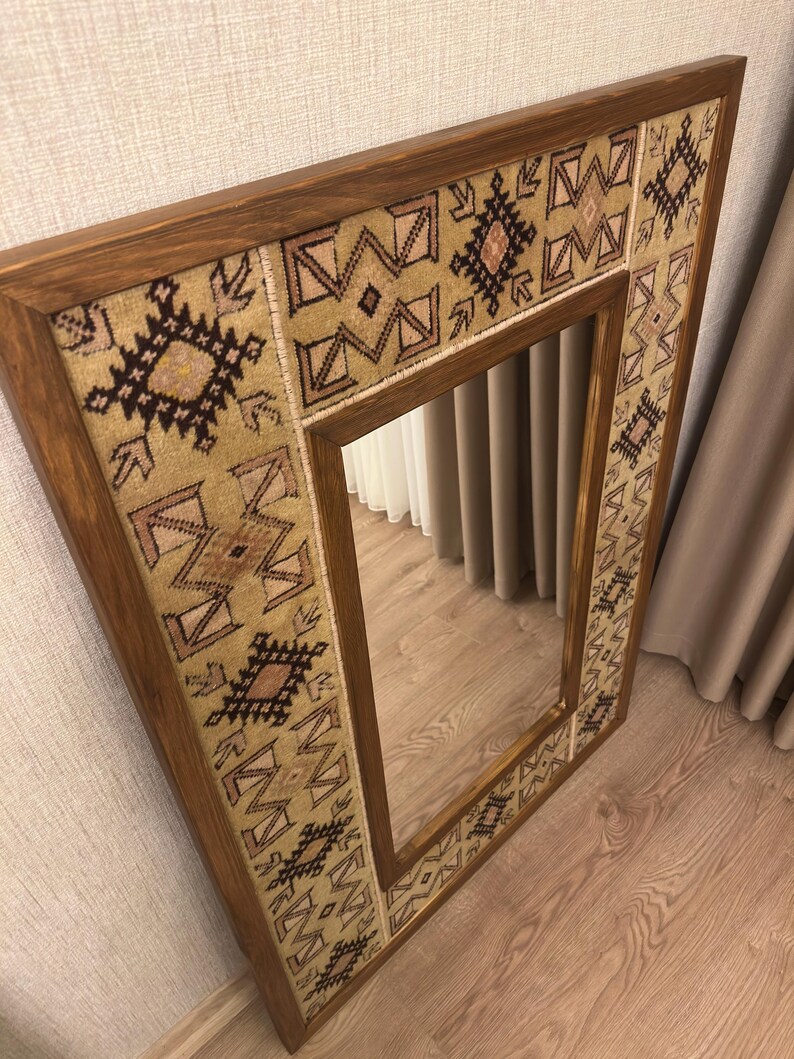 Puede incluir: Un espejo de pared rectangular con un marco de madera de color marr&oacute;n oscuro. El marco est&aacute; incrustado con un borde de tela beige texturizada, decorado con patrones geom&eacute;tricos repetitivos en tonos marr&oacute;n oscuro, negro y rosa suave.