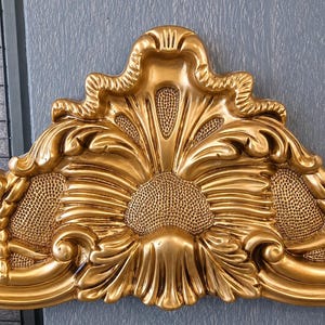 May include: A gold-colored decorative headboard crest featuring an ornate, symmetrical design with raised acanthus leaf carvings, scrolling edges, and textured, stippled recessed areas that create a high-contrast, three-dimensional appearance.