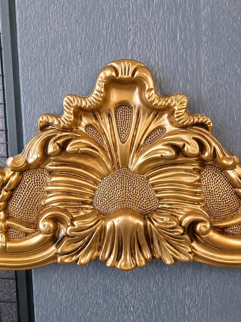 May include: A gold-colored decorative headboard crest featuring an ornate, symmetrical design with raised acanthus leaf carvings, scrolling edges, and textured, stippled recessed areas that create a high-contrast, three-dimensional appearance.