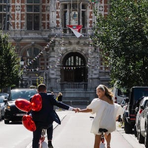 Op de afbeelding: Twee personen fietsen op een stadsstraat terwijl ze elkaars hand vasthouden. E&eacute;n persoon draagt een donkerblauw pak en heeft drie rode hartvormige ballonnen bij zich. De andere persoon draagt een korte witte jurk en een witte haarband. Een groot stenen gebouw staat op de achtergrond.
