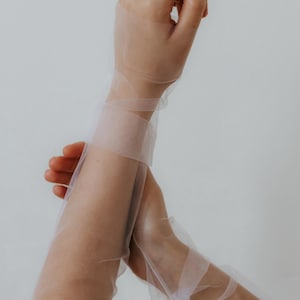 Puede incluir: Guantes largos de tul blanco transparente, recogidos en las muñecas con una cinta. Un delicado anillo adorna un dedo. Los guantes se extienden por los brazos. El fondo es blanco liso.