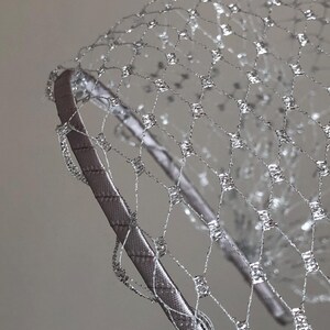 Può includere: Primo piano di un copricapo argentato con un design a rete. Il copricapo presenta una rete argentata attaccata a una fascia argentata. La rete ha un motivo a diamante con accenti argentati. La fascia è rivestita in tessuto argentato.