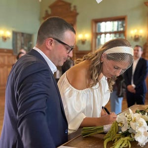 Op de afbeelding: Een persoon draagt een witte blouse met blote schouders en een witte haarband met een birdcage-sluier en ondertekent een document. Daarnaast ligt een boeket van witte orchidee&euml;n en groen amarant. Een persoon in een marineblauw colbert staat ernaast.