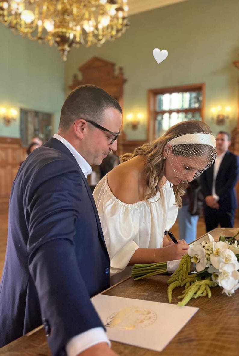 Op de afbeelding: Een persoon draagt een witte blouse met blote schouders en een witte haarband met een birdcage-sluier en ondertekent een document. Daarnaast ligt een boeket van witte orchidee&euml;n en groen amarant. Een persoon in een marineblauw colbert staat ernaast.